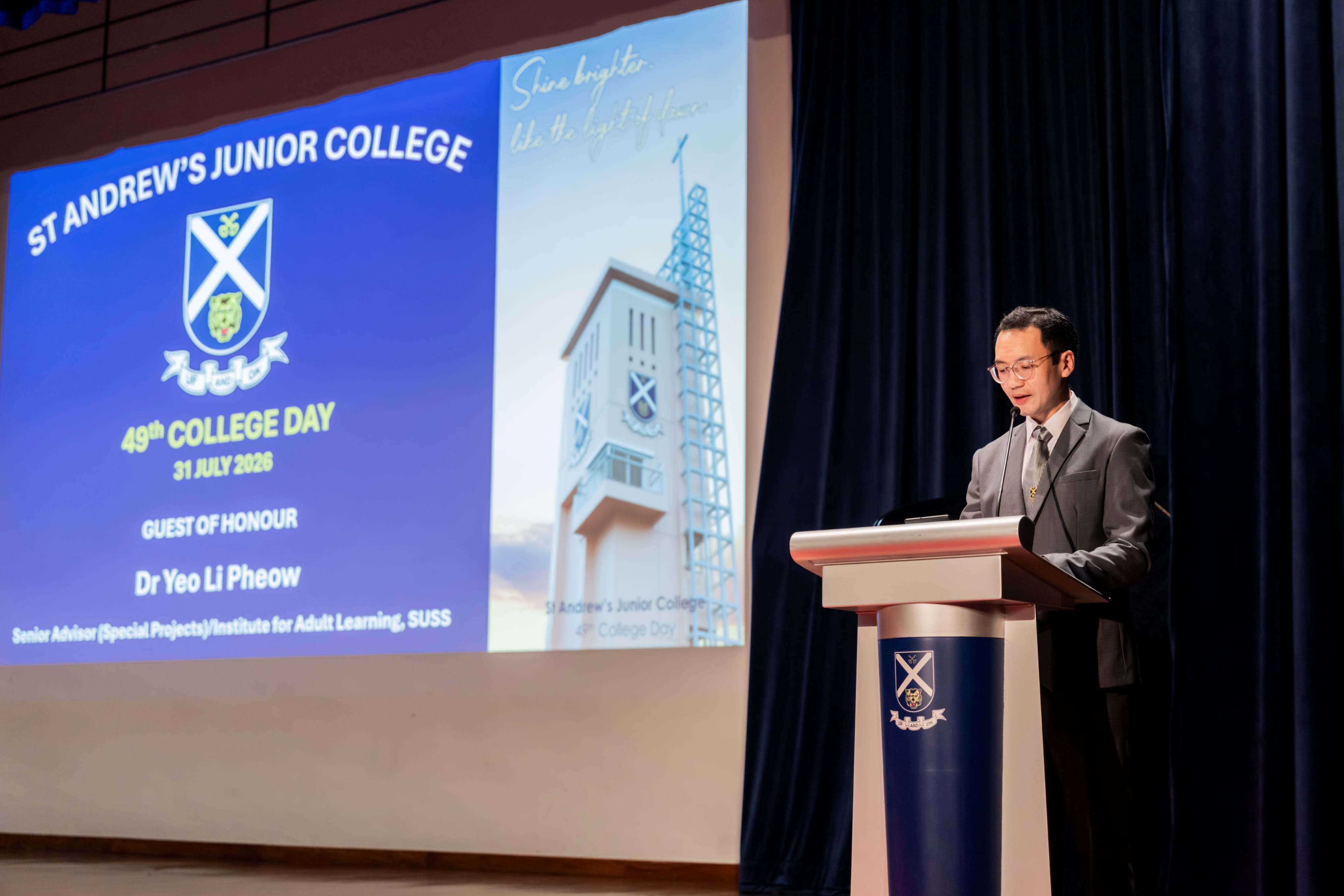 Mr Wayne Ko, emcee for the event, in a grey suit addresses the audience from behind a podium during the St Andrew's Junior College 49th College Day event, beside a large presentation screen.